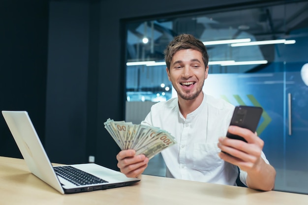 Foto o homem trabalha no escritório o empresário detém dólares em dinheiro se alegra e sorri olha para a tela do telefone se alegra com a vitória