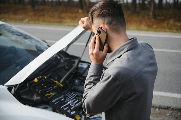 O homem usa uma garagem de chamada de celular em frente ao capô aberto de um carro quebrado na estrada na floresta Conceito de avaria do carro