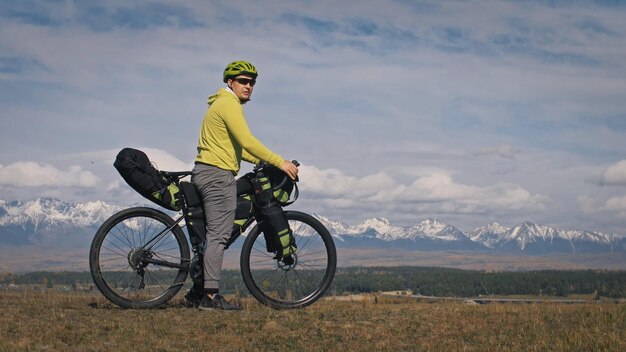 O homem viaja em terreno misto cicloturismo com bikepacking A viagem do viajante com malas de bicicleta Turismo esportivo bikepacking bike sportswear nas cores pretas verdes Montanhas cobertas de neve