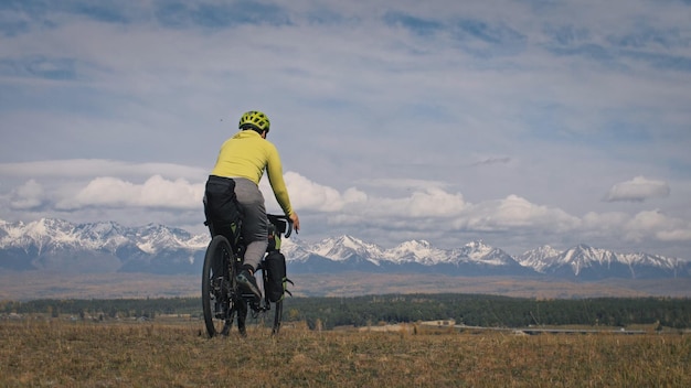 O homem viaja em terreno misto cicloturismo com bikepacking A viagem do viajante com malas de bicicleta Turismo esportivo bikepacking bike sportswear nas cores pretas verdes Montanhas cobertas de neve