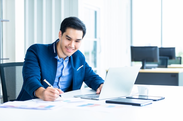 O humor feliz um alegre do jovem empresário asiático tem idéias para anotar o plano de negócios bem sucedido em papel de documento e laptop na mesa de madeira no escritório.