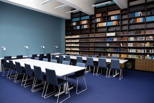 Foto o interior da biblioteca, cores azuis e marrons. estantes com livros, mesas brancas.