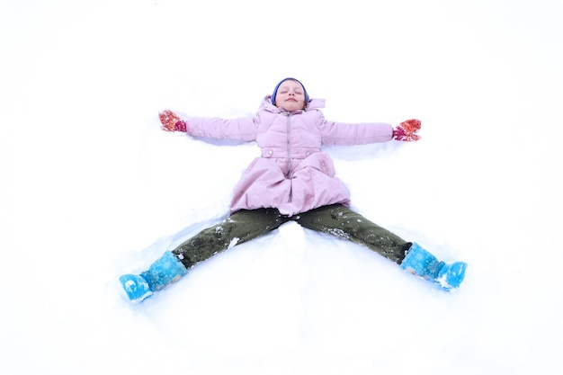 O inverno é uma época de jogo e prazer uma menina sorridente de oito anos em uma jaqueta pastel e um boné azul imita um anjo de neve em um quintal coberto de neve