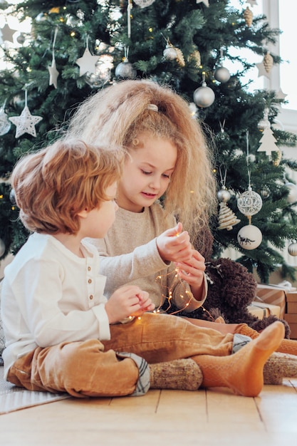O irmão e a irmã bonitos jogam com luzes e sentam-se sob a árvore de Natal