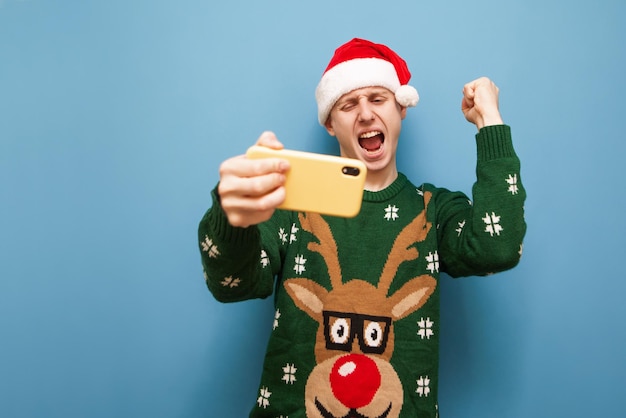 O jogador emocional em roupas de Natal jogando jogos para celular em um fundo azul se alegra