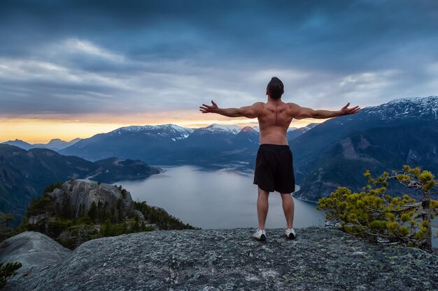 O jovem apto e musculoso está no topo da montanha durante um pôr do sol colorido