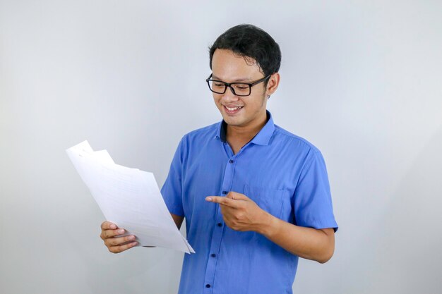 O jovem asiático está sorridente e feliz ao olhar para o documento em papel, indonésio vestindo camisa azul