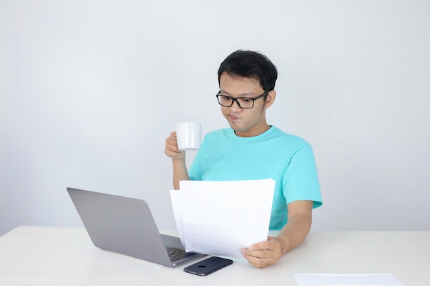 O jovem asiático está sorrindo e feliz ao trabalhar em um laptop e documento com caneca branca na mão indonésio vestindo camisa azul