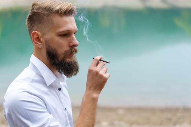 O jovem barbudo fuma cigarro ou o cara do tabaco é viciado em cigarros e sempre fuma muito ...