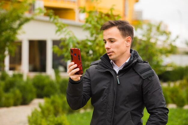 O jovem bonito está olhando para smartphone O homem vestindo uma jaqueta fica ao ar livre no outono