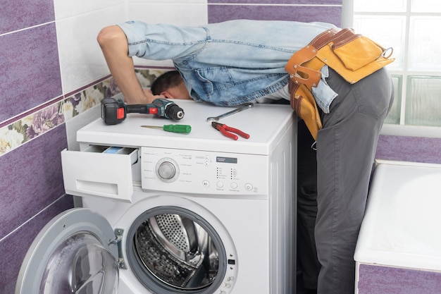 O jovem e bonito reparador em terno de trabalhador com a caixa de ferramentas profissionais está consertando a máquina de lavar no banheiro