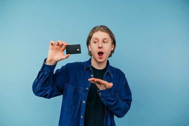 O jovem estudante positivo em uma camisa fica em um fundo azul com um cartão bancário na mão