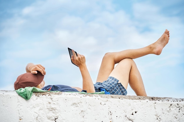 O jovem lê o livro digital no tablet deitado no cais de pedra com as pernas nuas contra o céu azul