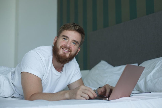 O jovem mente e usa o laptop na cama de manhã, conceito de aprendizado on-line, compras de trabalho