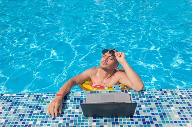 O jovem nas férias de verão com o laptop, negócios on-line. Escritório em casa à distância na piscina em anel de borracha em quarentena