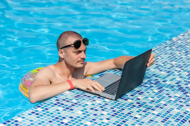 O jovem nas férias de verão com o laptop, negócios on-line. escritório em casa à distância na piscina em anel de borracha em quarentena