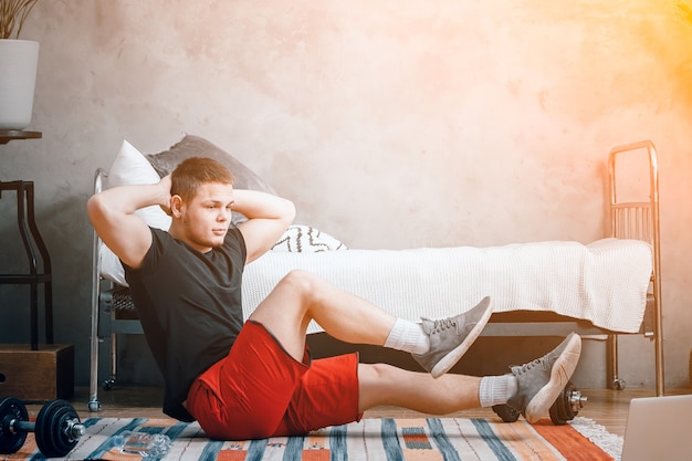 O jovem pratica esportes em casa. esportista alegre com cabelo preto faz a imprensa no quarto, ao lado há um laptop com treino online