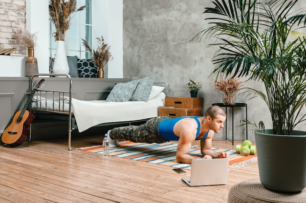 O jovem pratica esportes em casa prancha e assiste a exercícios on-line em um laptop no quarto