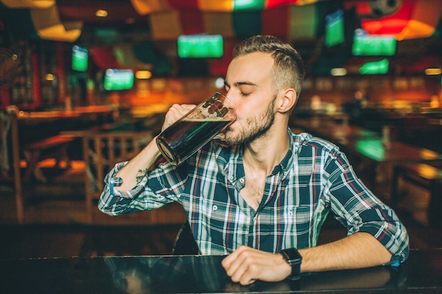 O jovem senta-se sozinho no balcão de bar no pub. Ele bebe cerveja escura da caneca.