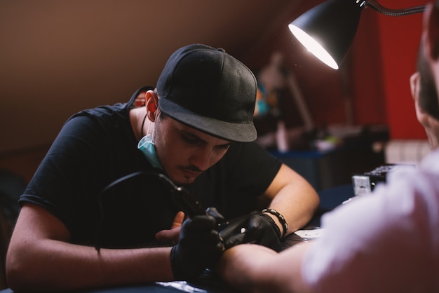 O jovem tatuador focado está pintando os braços dos clientes com cuidado em sua loja.