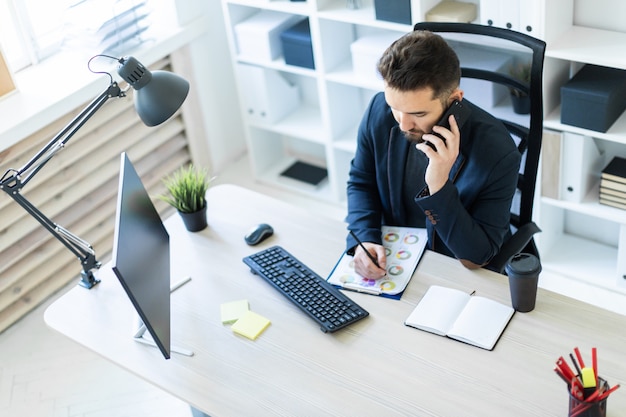 O jovem trabalha no escritório em uma mesa de computador com documentos, diagramas e telefone.