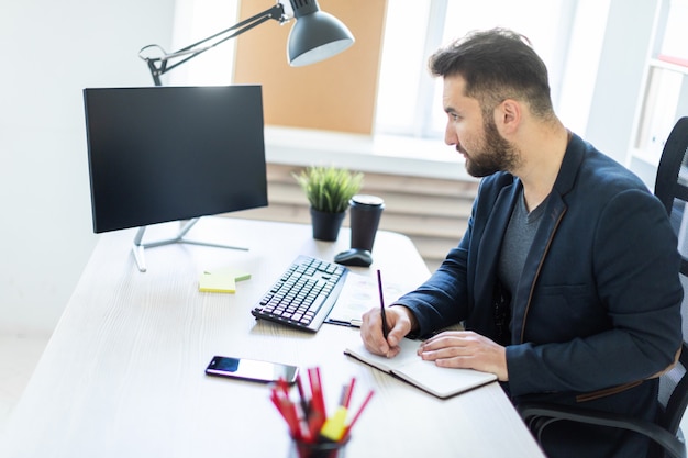 O jovem trabalha no escritório em uma mesa de computador com documentos, diagramas e telefone.