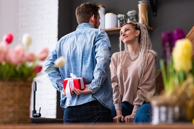 O jovem vai dar um lindo presente para uma garota A garota está sentada na cozinha o cara está segurando um presente nas costas Jovem casal alegre em roupas casuais