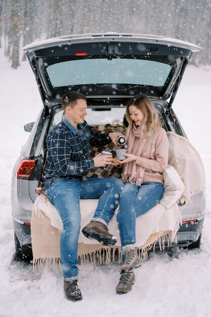 O marido despeja café de uma garrafa térmica na xícara de sua esposa enquanto está sentado com ela no porta-malas de um carro