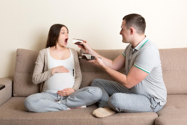 O marido feliz está alimentando sua esposa grávida com um pedaço de bolo relaxando no sofá em casa O futuro pai cuida da mãe grávida