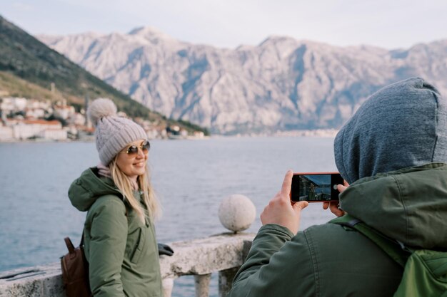 O marido fotografa sua esposa apoiada nas barragens na costa do mar com vista para as montanhas.