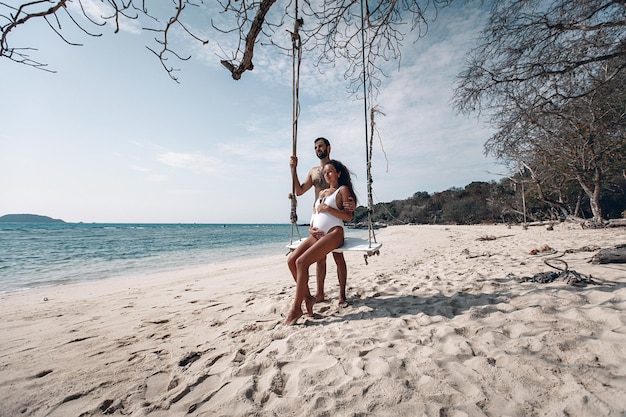 O marido orgulhoso abraça ternamente a esposa grávida, enquanto ela se senta nos balanços acariciando a barriga na praia branca junto às águas azuis; conceito de harmonia.