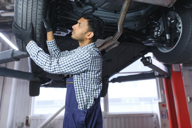 O mecânico de automóveis hispânico latino de uniforme está examinando um carro enquanto trabalha no serviço de automóveis