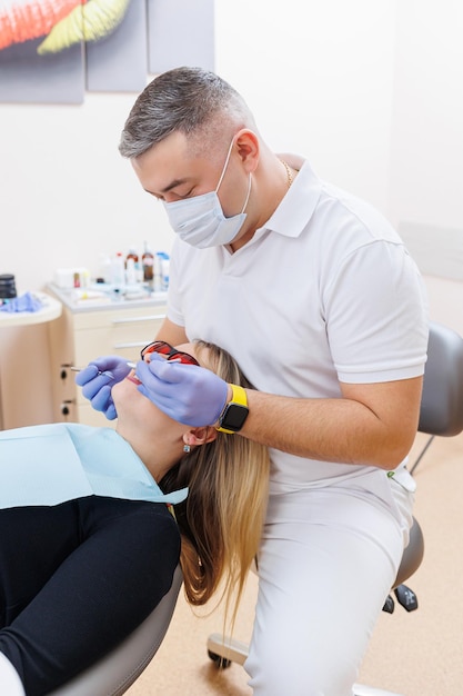 O médico dentista olha para os dentes do paciente e segura os instrumentos dentários perto da boca O assistente ajuda o médico Eles usam uniformes brancos com máscaras e luvas Dentista Consultório odontológico