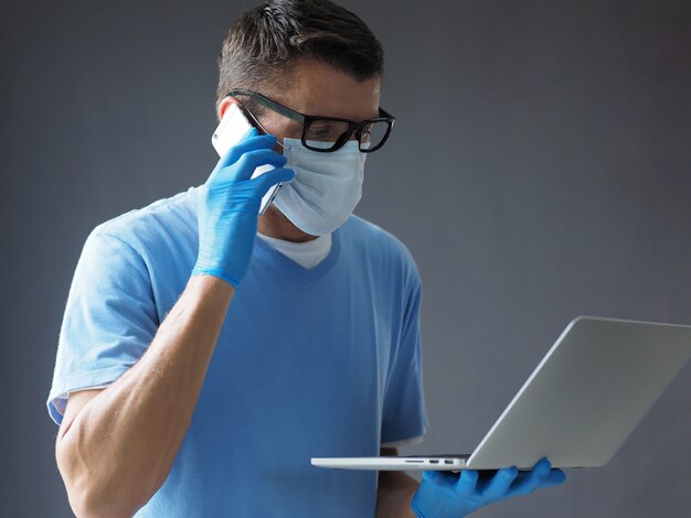 O médico homem usa máscara facial usando o celular e o laptop dele. Ocupado durante a epidemia de coronavírus.