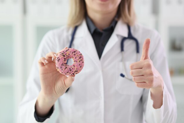 O médico segura rosquinha doce na mão mostrando o polegar para cima gesto na clínica mulher em uniforme médico