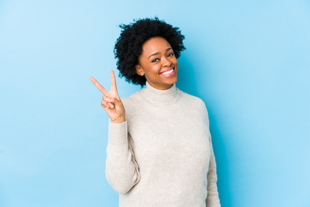 O meio envelheceu a mulher afro-americano contra uma parede azul isolada mostrando alegre e despreocupado um símbolo de paz com dedos.