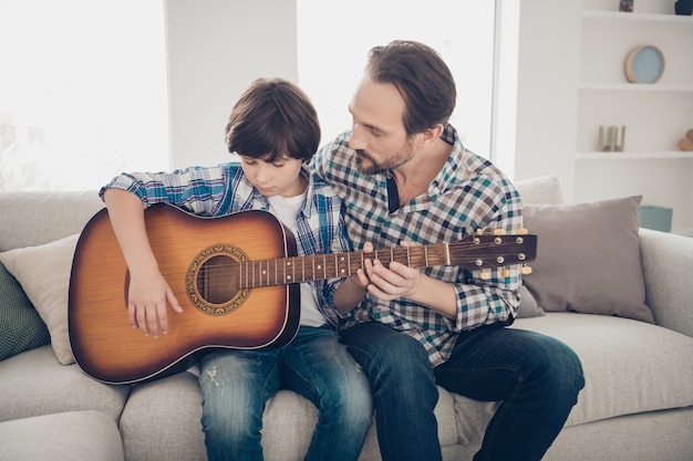 O melhor professor é o conceito de seus parentes. Pai maduro, concentrado e focado, ajudando o aluno a aprender a tocar guitarra segurando as mãos dos instrumentos, vestindo uma camisa xadrez casual sentado no sofá da casa