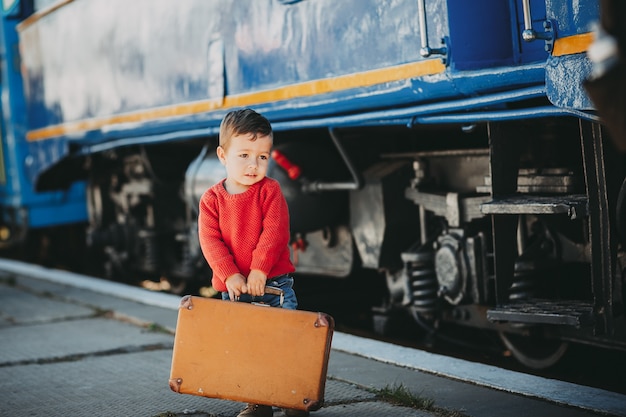 O menino adorável da criança vestiu-se na camisola vermelha em uma estação de trem perto do trem com a mala marrom velha retro.