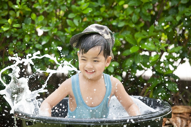 O menino asiático bonito se diverte jogando na água de uma mangueira em um dia de verão quente e ensolarado