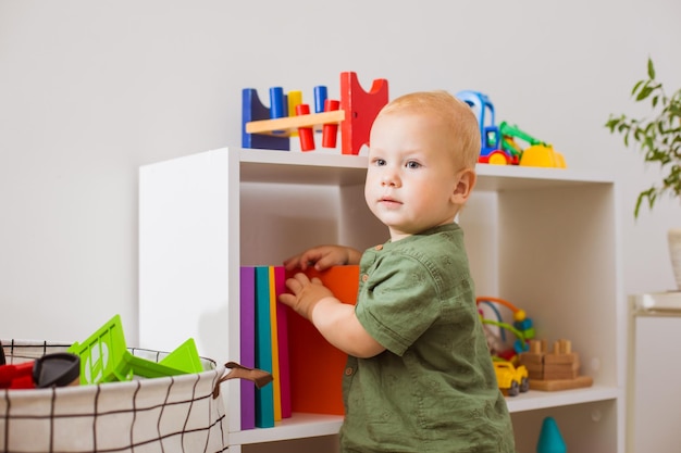 O menino bonitinho tira livros do armário e desvia o olhar O menino está de pé na sala de jogos perto da estante