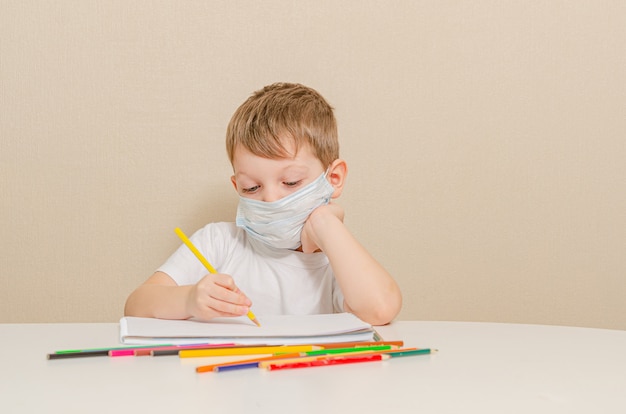 O menino bonito de 4 anos na máscara médica está sentado em casa em quarentena. entretenimento para a criança durante a quarentena - desenho.