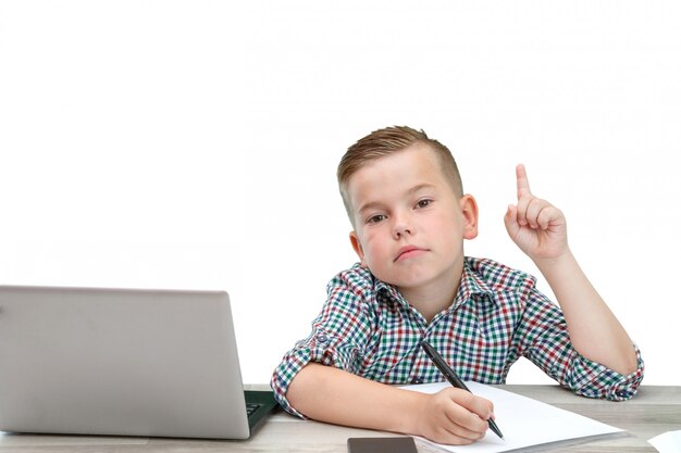 O menino caucasiano em idade escolar em uma camisa xadrez em um fundo isolado com um laptop e um telefone registra pensamentos em um pedaço de papel.