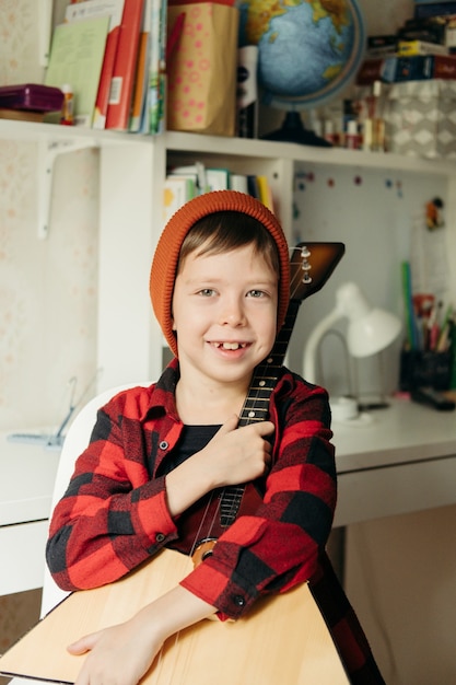 O menino com um chapéu vermelho e uma camisa xadrez joga a balalaica. rapaz bonito segurando seu violão. aulas de música em casa. hobby para a alma. música para ensino familiar