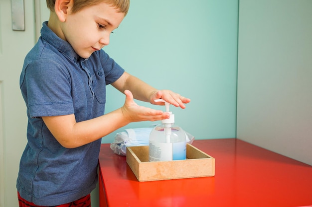 O menino criança usa anti-séptico no gel de mão do jardim de infância está em uma mesa vermelha