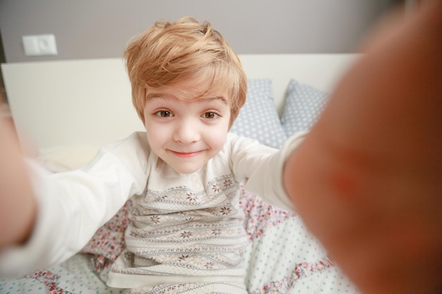 O menino de pijama em idade pré-escolar se senta na cama e se fotografa.