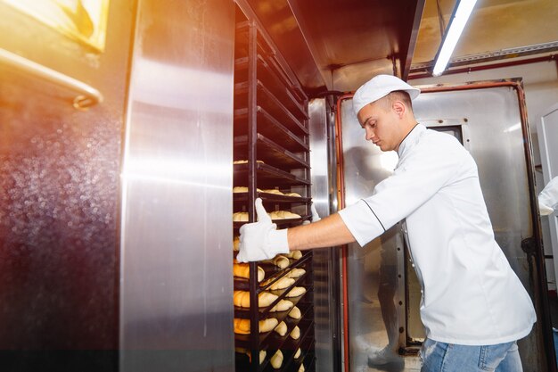 O menino do padeiro introduz um carro com bandejas de cozimento cruas da massa em um forno industrial em uma padaria.