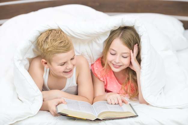 O menino e a menina felizes com um livro deitados sob o edredom