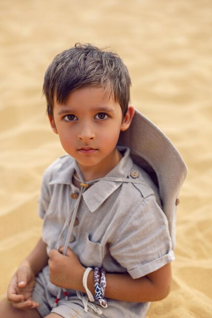 Foto o menino é uma criança que viaja com um terno de rastreador de arqueólogo e um chapéu sentado na areia do deserto