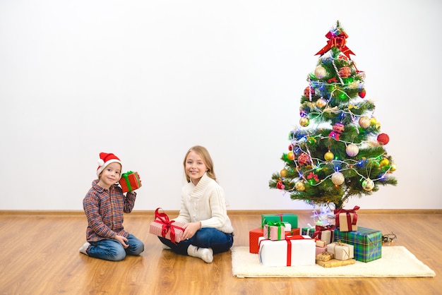 O menino e uma menina felizes sentam-se com caixas de presente perto da árvore de Natal
