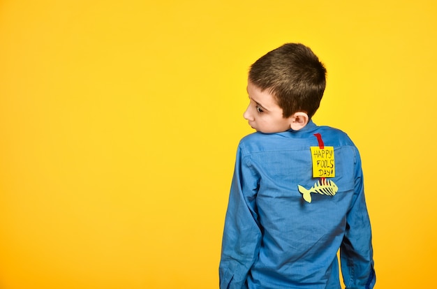 O menino em um fundo amarelo em uma camisa azul com uma fita colada de peixe e um pedaço de papel nas costas